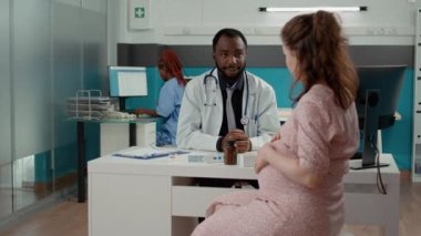 Erkek kadın doğum uzmanı hamile bir hastayla kontrol randevusu yapıyor. Tıp kabinesinde doğum ve doğum hakkında konuşuyor. Hamilelik göbeği olan sağlık uzmanı bir kadın..