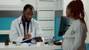 Hamilelik göbeği olan bir hastayla konuşan erkek doktor, dolap masasındaki kağıtlara not alıyor. Kadın doğum uzmanı. Çocuk bekleyen bir kadın. Muayene dosyalarına yazıyor..