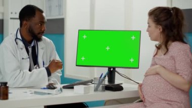 Hamilelik göbekli bir doktor ve hasta dolaptaki bilgisayardan yeşil ekrana bakıyor. Erkek doktor ve hamile kadın izole edilmiş kroma anahtar kalıplarını ve boş maketleri analiz ediyor. Üçayak atışı.