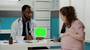 Doğum uzmanı tablet üzerinde yatay yeşil ekran ile çalışıyor ve gelecekteki annesiyle muayenede kullanıyor. İzole edilmiş telif alanı ve krom anahtarı olan boş model şablonu tutan erkek doktor.