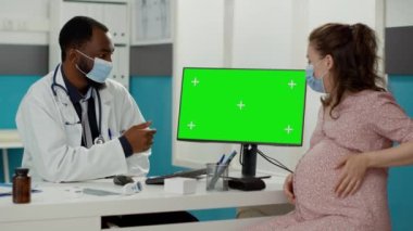 Hamilelik göbeği olan doktor ve hasta covid 19 salgını sırasında bilgisayarda yeşil ekrana bakıyor. Monitörde izole edilmiş krom anahtar şablonu ve boş kopya alanı.