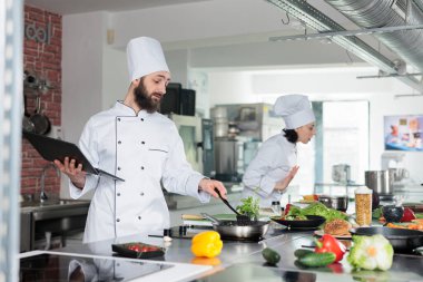 Gurme mutfak şefi yemek pişirirken bilgisayarla tarifi yakından takip ediyor. Gastronomi uzmanı mutfakta dizüstü bilgisayar tutarken taze organik malzemeler kullanarak yemek hazırlıyor..