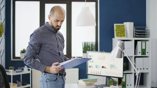 Ofis çalışanı araştırma belgelerindeki iş çizelgelerini analiz ediyor, satış istatistikleriyle finansal gelişimi planlamak için bilgisayar üzerinde çalışıyor. Erkek çalışan kağıtlardaki grafiklere bakıyor..