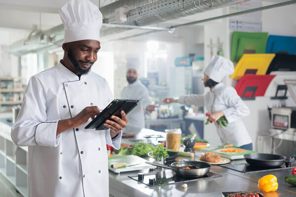 Gastronomy expert with handheld device standing in restaurant professional kitchen while looking for dinner recipes. Head cook with tablet brainstorming garnish ideas for gourmet cuisine meal.