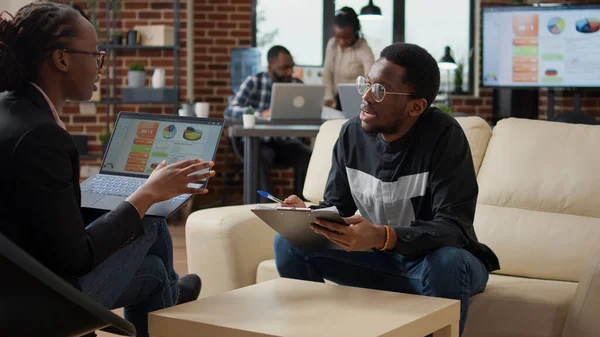 Woman explaining data charts on laptop and man taking notes on documents, doing job collaboration to create financial analysis strategy. Business people working together to plan project.