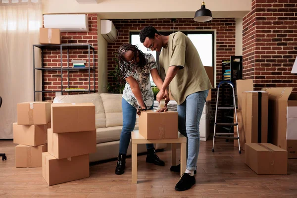 African american life partners packing up furniture in boxes using adhesive tape to move in new household property bought on loan. New beginnings in apartment, storage transportation.