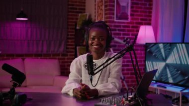 Afro-Amerikan Podcaster 'ın portresi evde kayıt stüdyosunda mikrofon ve dijital ses mikseriyle oturuyor. Çevrimiçi radyo sunucusu canlı yayın yaparken kameraya kendinden emin bir şekilde gülümsüyor.