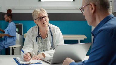 Sağlık uzmanı kabinedeki check-up muayenesinde notlar alıyor, randevuda kıdemli hastayla tartışıyor. Doktor, hastalıkları tedavi etmek için sağlık raporu yazıyor..