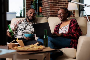 Kız arkadaş ve erkek arkadaş dizüstü bilgisayar kullanıyor ve paket yemek yiyor, televizyonda film izlemek için yemek dağıtımının keyfini çıkarıyor. Film izlemek, fast food ve birayla yemek yemek..