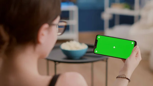 Closeup of young woman holding horizontal smartphone with green screen watching influencer vlog in living room. Girl using touchscreen mobile phone with chroma key looking at video content on screen.