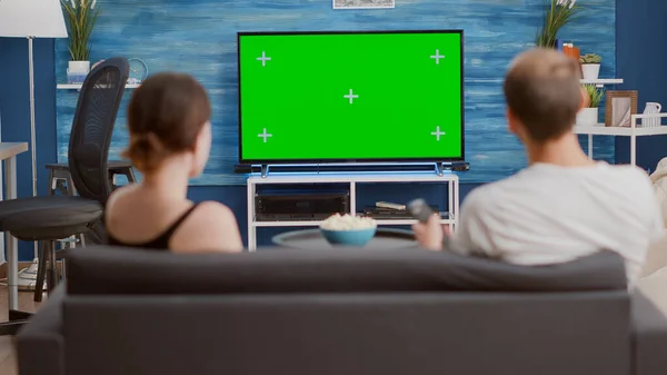 Back view of man browsing tv channels on green screen looking for girlfriend favourite show while sitting on couch. Couple enjoying television shows on chroma key mockup screen in modern living room.