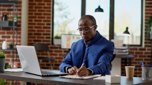 African american businessman taking notes on laptop, using internet statistics to create startup presentation. Company worker writing email, browsing website to plan project. Tripod shot.