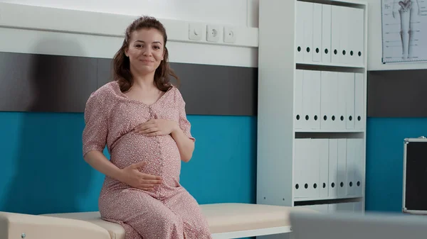 Portrait of future mother with baby bump sitting on cabinet bed, waiting to receive health care consultation from medic. Pregnant woman attending checkup appointment in office.