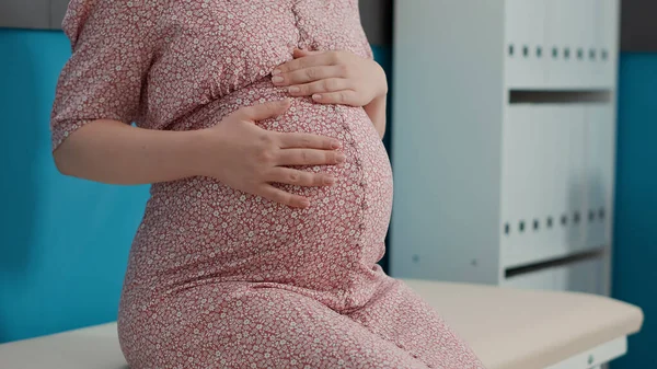 Pregnant patient attending checkup visit with obstetrician, sitting on bed in medical office. Woman with pregnancy belly preparing for childbirth, waiting on health specialist. Close up.