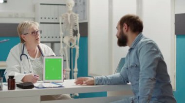 Physician and patient analyzing digital tablet with greenscreen in medical office. Man and medic using blank copyspace mockup with isolated chromakey background and template. Tripod shot.