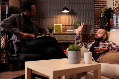 Erkek hasta psikologla terapi seansına katılıyor, kanepede dinleniyor ve davranış sorunları hakkında konuşuyor. Uzmanlara psikolojik sorunları açıklamak, profesyonel destek almak.