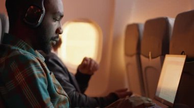 Uçuş sırasında dizüstü bilgisayar kullanarak iş seyahatinde uçan Afrika kökenli Amerikalı yolcu. Gün batımında ticari havayollarıyla seyahat etmek, havacılık jetinde kulaklık takmak. El kamerasıyla..
