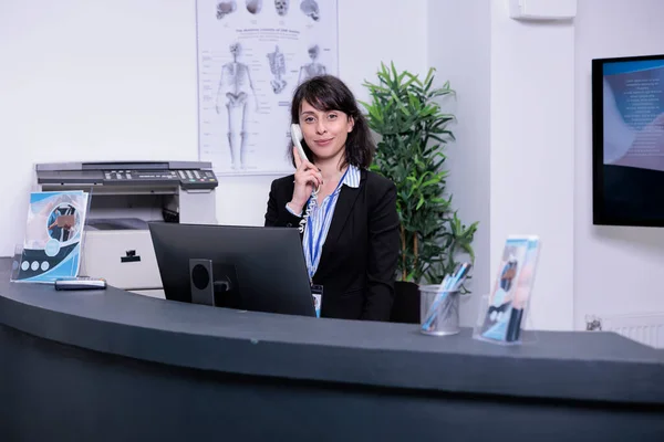 Portrait of smiling hospital receptionist answering call from patient to make an appointment for clinical consult at private clinic. Professional looking woman working at front desk using telephone.