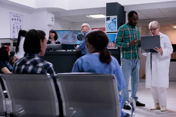 老年妇女医生在私人诊所候诊室拿着带有Mri扫描结果的笔记本电脑与非洲裔美国病人交谈 资深医生与较年轻的男子交谈 — 图库照片