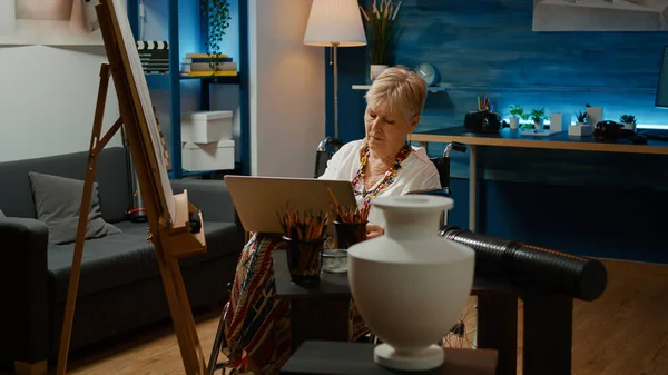 老年妇女长期受损使用笔记本电脑在白色帆布上画花瓶草图 残疾人士轮椅使用者与健康问题绘画大师 以电脑为灵感 — 图库照片