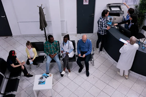 Top view of private clinic front desk with asian patient registering for doctor appointment. Diverse group of patients wait sitting in hospital lobby for clinical consult in hospital waiting area.