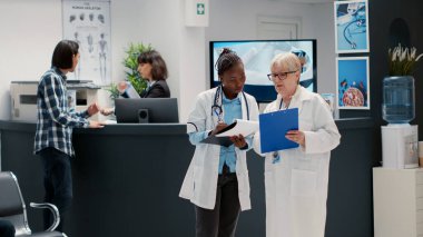 Kıdemli sıhhiyeci ve Afrikalı Amerikalı doktor teşhis koymak ve hastaları tedavi etmek için raporlara bakıyor, hastane resepsiyonunda konuşuyorlar. Bekleme alanında çok ırklı tıbbi personel var. Üçayak atışı.