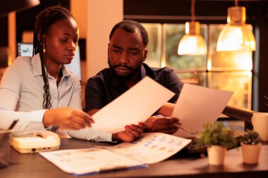 İş ortaklarından oluşan bir ekip rapor hazırlamak için beyin fırtınası yapıyor, belgelerle ilgili verilerle proje araştırması yapıyor. Gün batımında büyük pencereli sunum yapmak için evraklar analiz ediliyor.