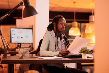 Genç bir çalışan veri tablolarıyla iş evraklarını analiz ediyor, bilgisayarda sunum yapıyor. Gün batımında şirket raporu üzerinde çalışmak için araştırma bilgilerine sahip belgelere bakıyorum..