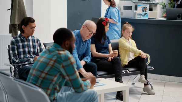 Multiethnic people waiting in hospital reception lobby to attend checkup appointment and talk to doctor. Diverse patients sitting in waiting area at facility, medical insurance support.