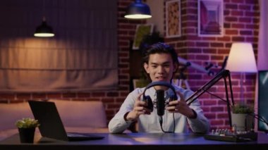 Male vlogger recording product review video with headphones, recommending wireless headset on camera. Creating channel content with modern earphones gadget on podcast episode.