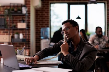 Fiziksel engelli bir şirket çalışanı sabit hattan konuşuyor, ofis telefonunu kullanıyor. Tekerlekli sandalye kullanıcısı çevrimiçi uzaktan konuşma yapıyor, engelle uğraşıyor.