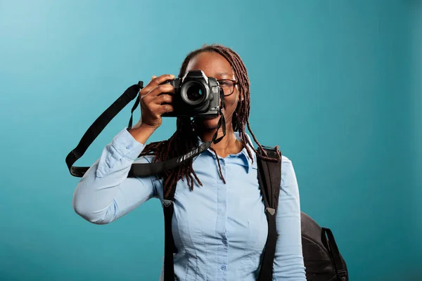 Confident and creative photographer taking pictures while enjoying leisure time on blue background. Young adult photography entusiast with modern photographing device taking photos. Studio shot