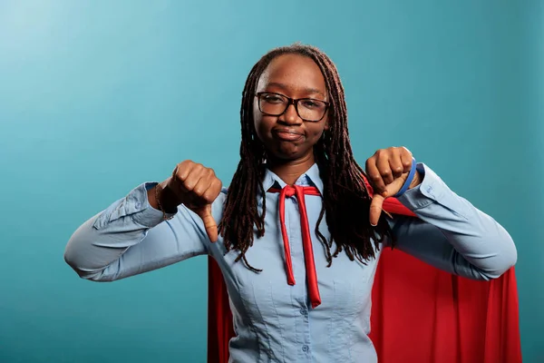 Dissapointed young adult superhero woman giving thumbs down dissaproval fingers gesture while looking at camera. African american mighty justice defender showing disagree hand symbol in disagreement.