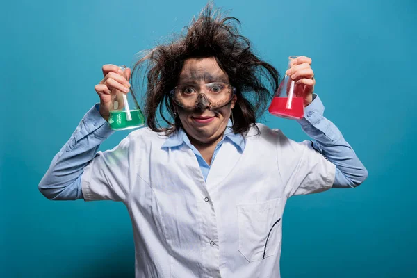 Goofy looking wacky scientist with messy hairstyle and dirty face holding professional glass jars filled with toxic chemical compounds. Mad amusing chemist having beakers filled with liquid substances