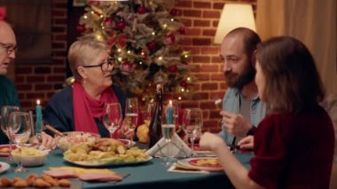 Yaşlı çift, Noel yemeğinde geleneksel ev yemeği yerken evli genç yetişkinlerle konuşuyor. Şenlikli insanlar kış tatilini otantik yemek ve şampanyayla kutluyor..