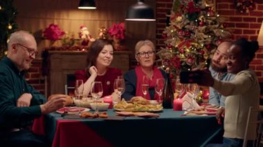 Cheerful family taking selfie photos with smartphone while sitting at Christmas dinner table. Festive diverse people celebrating winter holiday together inside warm apartment with Xmas decorations.