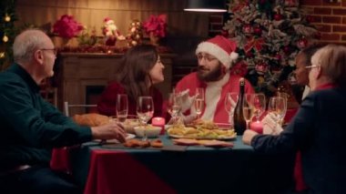 Funny person costumed as Santa Claus conversating with family about gifts while enjoying Christmas dinner at home. Festive man with traditional costume celebrating winter holiday with relatives.