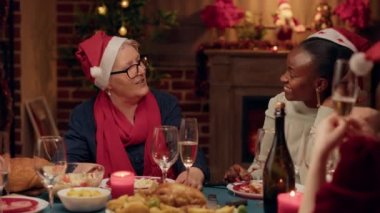 Kıdemli kadın evde Noel yemeğinin tadını çıkarırken neşeli, genç bir yetişkinle konuşuyor. Kış tatilini birlikte kutlarken şampanya içen çok kültürlü insanlar..