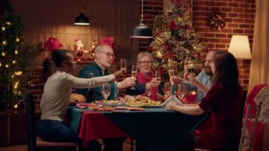 Cheerful multiethnic group of people celebrating winter holiday with sparkling wine. Happy close family members clinking champagne glasses while sitting together at Christmas dinner table. Tripod shot
