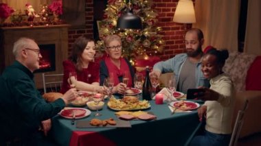 Beautiful african american woman with smartphone taking selfie photo of family celebrating winter holiday at home. Joyful diverse people enjoying Christmas dinner while creating memories.