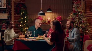 Positive multicultural family members celebrating Christmas with special evening dinner at home. Happy people sitting inside warm and cozy apartment decorated with authentic Xmas garlands. Tripod shot