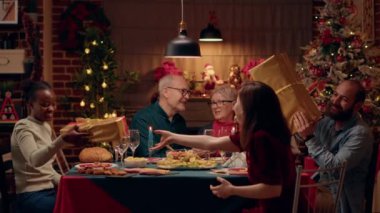 Festive family members sitting at Christmas dinner table while giving each other presents. Happy multiethnic people celebrating winter feast with gifts and traditional home cooked food. Handheld shot