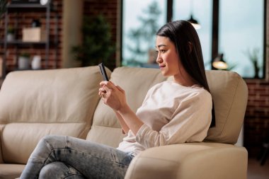 Ev telefonu teknolojisi kullanan güzel Asyalı kadın, internet kullanıcısı. Kanepede oturan mutlu, genç bir bayan. Elinde akıllı telefonuyla.