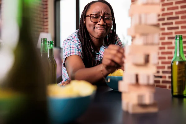 Smiling woman sitting at table in living room while playing society games with friends. Joyful person pulling wooden block from wooden tower while sitting at home and enjoying fun leisure activity.