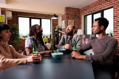 Birlikte alkollü içecekler içerken ve yeniden bir araya gelişimizi kutlarken masada oturan mutlu genç yetişkinler. Evde otururken bira ve atıştırmalıkların tadını çıkaran çok ırklı bir arkadaş grubu..