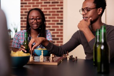 Düşünceli Afro-Amerikalı arkadaşlar oturma odasında oturmuş satranç oynuyorlar. Neşeli akıllı insanlar evlerinde satranç oynayarak rahatlarken abur cubur ve içeceklerin tadını çıkarıyorlar..