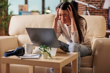 Genç Asyalı kadın evde çalışırken migren ve baş ağrısı çekiyor. Kanepede oturan yorgun kadın alnını örtüyor ve yorgun kırmızı gözler mutsuz, hastalıklı, bıkkın, bıkkın.