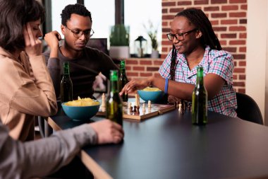 Akıllı insanlar birlikte satranç oynarken oturma odasında otururlar. Mutlu çoklu etnik arkadaşlar evlerinde oturup strateji oyunları, abur cuburlar ve içeceklerle eğleniyorlar..