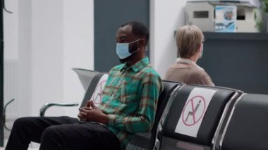 Young man waiting in hospital reception lobby to start consultation with medic during coronavirus pandemic. Diverse people sitting in waiting area chairs to do covid 19 consultation.