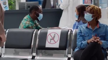 Multiethnic group of people with face masks sitting in waiting area at hospital reception lobby. Nurse and african american man talking about healthcare service during covid 19 epidemic.